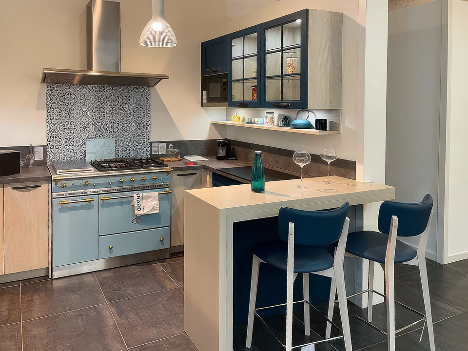 Façade moderne d'un showroom d'aménagement intérieur situé dans une zone commerciale, avec une large devanture vitrée illuminée présentant des aménagements de cuisine et salle de bain en vitrine, parking accessible devant le bâtiment, carrefour routier visible indiquant une bonne accessibilité routière, clients se dirigeant vers l'entrée du magasin en journée. Photo prise de face, mettant en avant la façade accueillante et la localisation dans une zone facilement accessible.