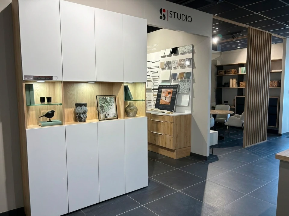Façade moderne d'un showroom d'aménagement intérieur situé dans une zone commerciale, avec une large devanture vitrée illuminée présentant des aménagements de cuisine et salle de bain en vitrine, parking accessible devant le bâtiment, carrefour routier visible indiquant une bonne accessibilité routière, clients se dirigeant vers l'entrée du magasin en journée. Photo prise de face, mettant en avant la façade accueillante et la localisation dans une zone facilement accessible.